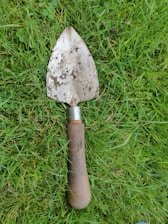 A trowel with a well-worn wooden handle and a metal blade covered in soil rests on lush green grass. The grass is slightly uneven, with a mix of small clover leaves and blades. The trowel appears aged and used, indicating its frequent use in gardening or landscaping tasks.
