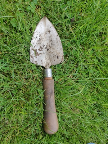 A trowel with a well-worn wooden handle and a metal blade covered in soil rests on lush green grass. The grass is slightly uneven, with a mix of small clover leaves and blades. The trowel appears aged and used, indicating its frequent use in gardening or landscaping tasks.