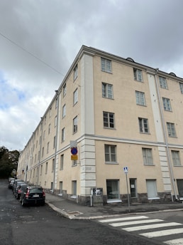 A multi-story beige residential building with multiple windows, situated at the corner of a street. The structure has a simple architectural design with white accents around the corners and windows. Several cars are parked on the street alongside the building. A street sign near the corner indicates parking regulations, and a pedestrian crossing is visible in the foreground. The sky is overcast, contributing to a somewhat muted lighting over the scene.