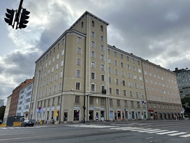 A tall beige building with multiple windows standing on the corner of a busy intersection. The ground floor has several storefronts, and there are Finnish flags attached to the building. Overcast skies and traffic lights are visible, with a zebra crossing in the foreground.