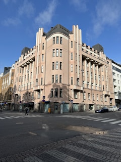 A large, multi-story building with an art deco design featuring rounded corners and symmetrical architecture. It is painted in light pink with multiple windows and black rooftop elements. The building is situated on a street corner, with cobblestone paving in the foreground. Pedestrians and cars can be seen on the road nearby.