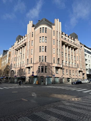 A large, multi-story building with an art deco design featuring rounded corners and symmetrical architecture. It is painted in light pink with multiple windows and black rooftop elements. The building is situated on a street corner, with cobblestone paving in the foreground. Pedestrians and cars can be seen on the road nearby.