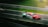 A racing car speeds down a track, featuring a dynamic motion blur in the background. The vehicle is prominently colored in red and green with a distinct number 60 on its body. The track is lined with metallic barriers and the grass is visible alongside the track.