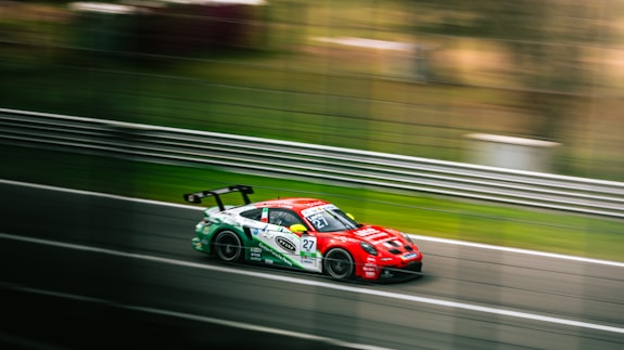 A sleek, red racing car speeds along a track, creating a sense of motion with the blurred background. The car features several sponsor logos and has a prominent rear wing. Metal guardrails line the track, and the environment is grassy, suggesting a professional racing event.