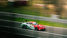 A sleek, red racing car speeds along a track, creating a sense of motion with the blurred background. The car features several sponsor logos and has a prominent rear wing. Metal guardrails line the track, and the environment is grassy, suggesting a professional racing event.