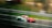A sleek, red racing car speeds along a track, creating a sense of motion with the blurred background. The car features several sponsor logos and has a prominent rear wing. Metal guardrails line the track, and the environment is grassy, suggesting a professional racing event.