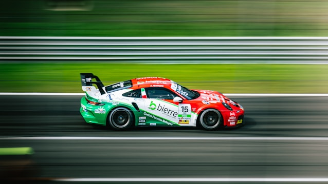 A race car with a vibrant red, white, and green design speeds along a track, creating a motion blur effect. The background consists of blurred green and gray lines, indicating high speed.