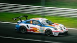 A colorful racing car featuring vibrant stripes of blue, red, and yellow on a white body is speeding on a racetrack. The car has number 8 and various sponsor logos, including 'Boss' and 'Sparco.' The background shows blurred greenery and a metal barrier, indicating high speed.