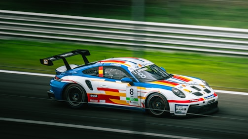 A colorful racing car featuring vibrant stripes of blue, red, and yellow on a white body is speeding on a racetrack. The car has number 8 and various sponsor logos, including 'Boss' and 'Sparco.' The background shows blurred greenery and a metal barrier, indicating high speed.