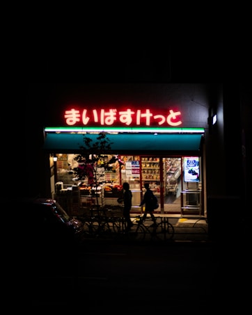 A dimly lit convenience store at night with a bright neon sign displaying Japanese characters above the entrance. The storefront is illuminated, revealing shelves stocked with various products, and two silhouetted figures walking past along with parked bicycles.