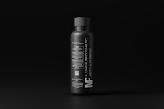 A sleek, matte black aluminum bottle stands against a dark background. The label features modern, minimalist typography in white, detailing the product as a cosmetic bottle mockup. The overall design conveys a luxurious and contemporary feel.