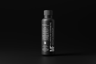 A sleek, matte black aluminum bottle stands against a dark background. The label features modern, minimalist typography in white, detailing the product as a cosmetic bottle mockup. The overall design conveys a luxurious and contemporary feel.