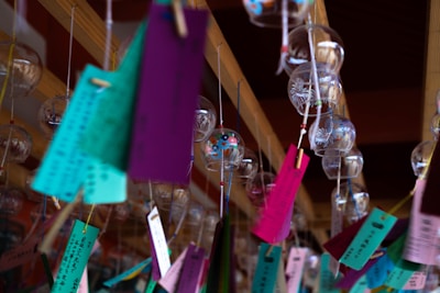 A series of colorful wind chimes hang from a wooden frame. Each chime is made of glass and decorated with intricate designs. Attached to the chimes are vibrant rectangular pieces of paper in various colors such as purple, green, and pink. The scene suggests a traditional or cultural setting.