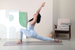 A person is performing a yoga pose on a mat in a minimalistic room. The individual is in a deep lunge with arms stretched upwards, demonstrating focus and balance. The background features abstract artwork and a chair with patterned cushions.