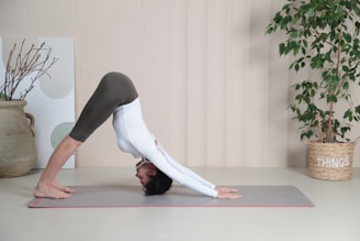 A person is performing a yoga pose resembling Downward Dog on a yoga mat in a well-lit room. The individual is wearing a long-sleeve white top and olive green leggings. A large potted plant and a basket labeled 'THINGS' are visible in the background, alongside a decorative pot with twigs and a canvas with abstract designs on the wall.