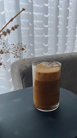 A glass filled with frothy coffee is placed on a dark tabletop. The background features a soft, light-colored chair and translucent curtains, creating a cozy and relaxed atmosphere. A sprig of dried flowers is positioned on the left side, adding a touch of natural decor.