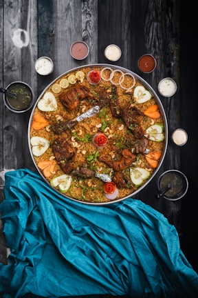 A large platter filled with spiced rice and several pieces of grilled meat. The dish is garnished with fresh herbs, cucumber slices, carrot wedges, and lemon slices. Small bowls of various sauces and dips surround the platter. The setup is on a dark, textured wooden surface, with a vibrant blue cloth.