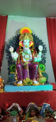 A statue of Lord Ganesha is seated on an ornate throne, surrounded by decorative elements. The deity has four arms, each holding different objects, symbolizing various divine attributes. The statue is adorned with a crown, floral garlands, and a colorful drape. Offerings, including bananas and coconuts, are placed at the base of the statue, surrounded by vibrant red drapery.