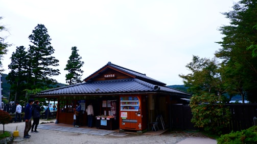 A traditional Japanese building with a tiled roof stands surrounded by trees. People are gathered near the entrance, where a vending machine is visible, and signs or posters are displayed. The area is bordered by greenery, and the sky is overcast.