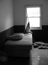 A small, dimly lit room contains a bare mattress with a single pillow placed on top. The floor appears worn, and there is a disheveled blanket or piece of fabric on the floor beside the mattress. A partially covered window allows some light into the room, casting shadows.