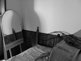 A small, cluttered room features a single bed with an unkempt mattress and a thick blanket draped over it. A freestanding oval mirror leans against the corner of the room, reflecting part of the bed frame. The walls are plain, and a wooden panel runs along the lower half of the walls.