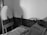 A small, cluttered room features a single bed with an unkempt mattress and a thick blanket draped over it. A freestanding oval mirror leans against the corner of the room, reflecting part of the bed frame. The walls are plain, and a wooden panel runs along the lower half of the walls.