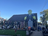 A house is decorated with various Halloween-themed items, including a large inflatable Frankenstein monster, several skeleton figures, and pumpkins. The yard is filled with numerous spooky decorations such as ghosts and witches, set against a clear blue sky.