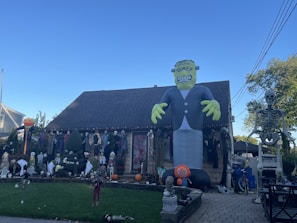A house is decorated with various Halloween-themed items, including a large inflatable Frankenstein monster, several skeleton figures, and pumpkins. The yard is filled with numerous spooky decorations such as ghosts and witches, set against a clear blue sky.