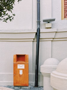 An orange mailbox is positioned against a light-colored, textured wall. The mailbox features an envelope symbol and some printed text. Above it, a black vertical pipe runs from the ground to the top of the image. Beside the pipe, there is a small, rectangular light fixture. Partially visible at the bottom right is a round white structure, possibly an architectural feature. A leafy branch protrudes from the upper left corner.