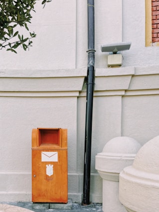 An orange mailbox is positioned against a light-colored, textured wall. The mailbox features an envelope symbol and some printed text. Above it, a black vertical pipe runs from the ground to the top of the image. Beside the pipe, there is a small, rectangular light fixture. Partially visible at the bottom right is a round white structure, possibly an architectural feature. A leafy branch protrudes from the upper left corner.