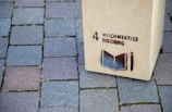 A wooden structure stands on a cobblestone pavement with the text '4 Hochwertige Bildung' and an image of an open book and pencil stenciled on it.