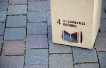 A wooden structure stands on a cobblestone pavement with the text '4 Hochwertige Bildung' and an image of an open book and pencil stenciled on it.
