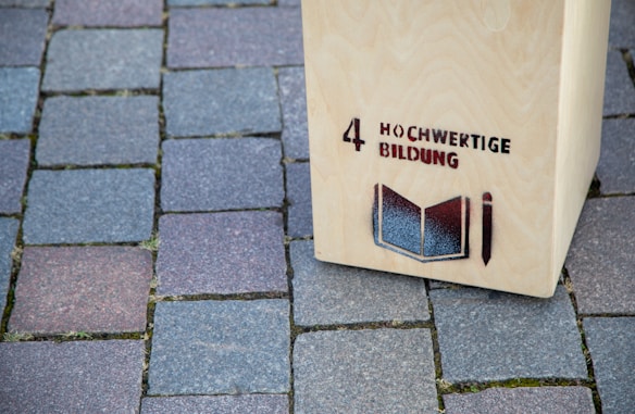 A wooden structure stands on a cobblestone pavement with the text '4 Hochwertige Bildung' and an image of an open book and pencil stenciled on it.