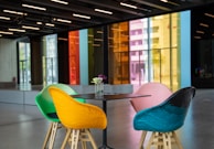 A modern interior space featuring a set of four colorful chairs around a minimalist black table. The chairs are vibrantly colored in green, yellow, pink, and blue. A small vase with flowers is placed on the table. In the background, large floor-to-ceiling windows with brightly tinted glass panels in red, orange, yellow, and blue provide a striking visual contrast. The room appears spacious and well-lit, with a polished concrete floor and overhead lighting.