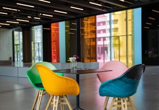 A modern interior space featuring a set of four colorful chairs around a minimalist black table. The chairs are vibrantly colored in green, yellow, pink, and blue. A small vase with flowers is placed on the table. In the background, large floor-to-ceiling windows with brightly tinted glass panels in red, orange, yellow, and blue provide a striking visual contrast. The room appears spacious and well-lit, with a polished concrete floor and overhead lighting.