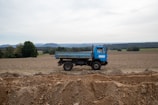 A blue dump truck is parked in an open, plowed field. The background features rolling hills and a line of trees under a cloudy sky. The earth in the foreground is disturbed, with mounds and trenches of dirt visible.