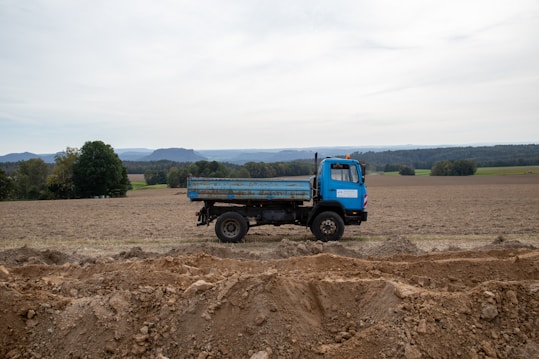 A blue dump truck is parked in an open, plowed field. The background features rolling hills and a line of trees under a cloudy sky. The earth in the foreground is disturbed, with mounds and trenches of dirt visible.