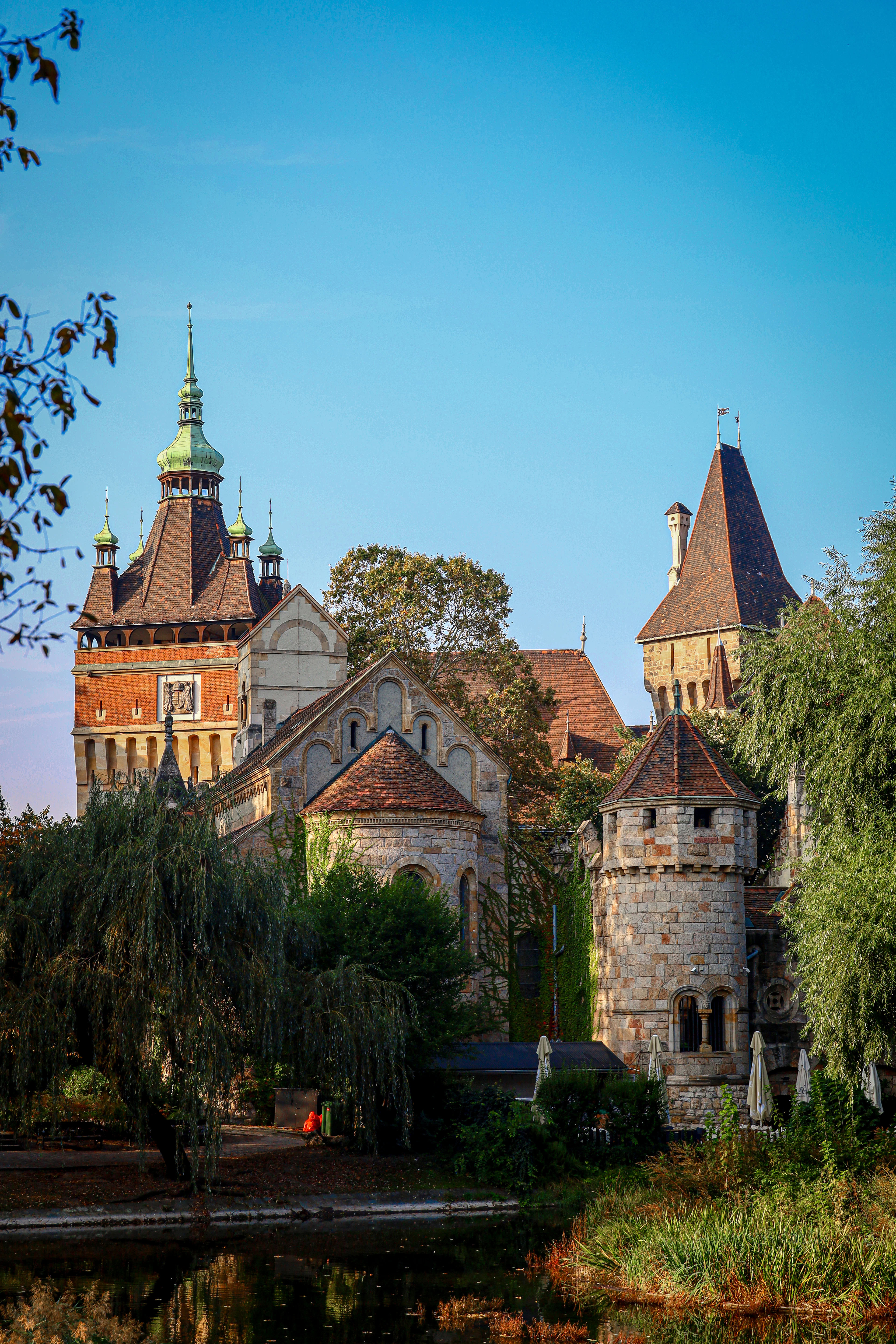 Vajdahunyad Castle in Budapest's City Park at golden hour, its green-capped spire, red roofs and round stone tower rising above willows and the still water of the park lake