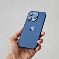 A hand is holding a sleek smartphone with a blue back featuring a prominent brand logo. The phone has a triple camera setup with a flash, positioned in an upper corner.