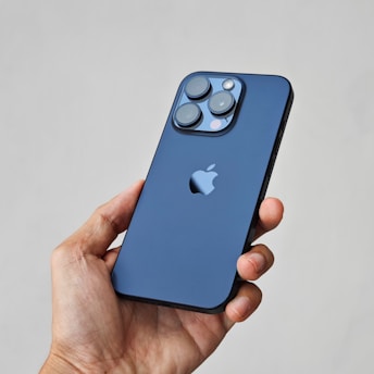 A hand is holding a sleek smartphone with a blue back featuring a prominent brand logo. The phone has a triple camera setup with a flash, positioned in an upper corner.
