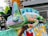 A colorful sculpture of a chameleon is perched on a decorative surface, surrounded by vibrant plants and urban elements in the background. The chameleon features a striking array of colors, including shades of green, blue, orange, and purple, blending seamlessly into the artistic surroundings.