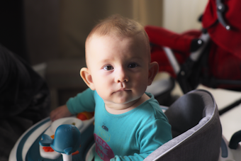 A baby with light hair and blue eyes is sitting in a baby chair. The baby is wearing a turquoise shirt, and there is a toy with a blue whale in front of them. In the background, there is a red stroller and partially visible furniture.