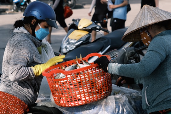 Two individuals are engaged in a street market transaction involving fish. One person, wearing a helmet, face mask, and gloves, appears to be selecting fish from a large red plastic basket held by another person. The second person is wearing a traditional conical hat and also has a face mask covering their mouth. In the background are parked motorbikes and other people walking, suggesting a bustling outdoor market setting.