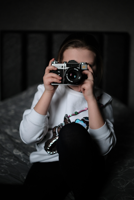 A young child is sitting on a bed holding a camera up to their face, taking a picture. The child is wearing a white sweatshirt with a colorful design and black pants. The background is out of focus, giving prominence to the child and the camera.