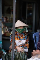 A person wearing a traditional conical hat and a colorful patterned garment sits in an outdoor setting. Their face is partially covered with a green patterned cloth. The surroundings include a blue plastic chair and a variety of goods and objects, suggesting a market environment. There are wrapped items resembling food products in the foreground.