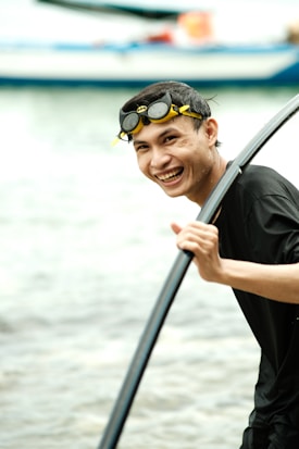 A person with short dark hair is holding a black paddle in their right hand while smiling joyfully. They are wearing dark swim goggles with yellow accents, standing near what appears to be a calm water body with boats visible in the background.