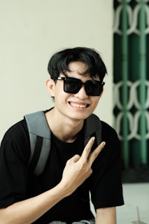 A person wearing sunglasses and a black shirt is posing with a peace sign gesture. They have a gray backpack slung over their shoulder. The background features a green and white geometric pattern.