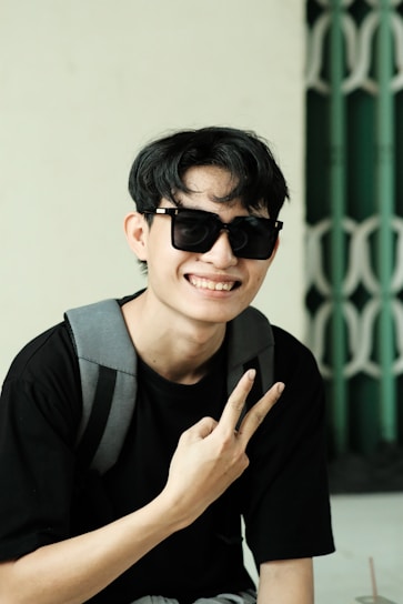 A person wearing sunglasses and a black shirt is posing with a peace sign gesture. They have a gray backpack slung over their shoulder. The background features a green and white geometric pattern.