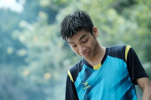 A person with wet hair is wearing a blue and black sports jersey, standing outdoors with a blurred green and yellow background. The person's facial expression appears curious or amused.