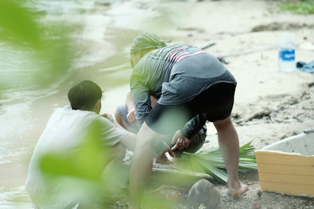 Three individuals are engaged in an outdoor activity near a body of water. They appear to be working together on something, possibly tied into some foliage or plant material. The environment suggests a natural, outdoor setting with sand, water, and some dense greenery in the background. A plastic water container is visible on the right.
